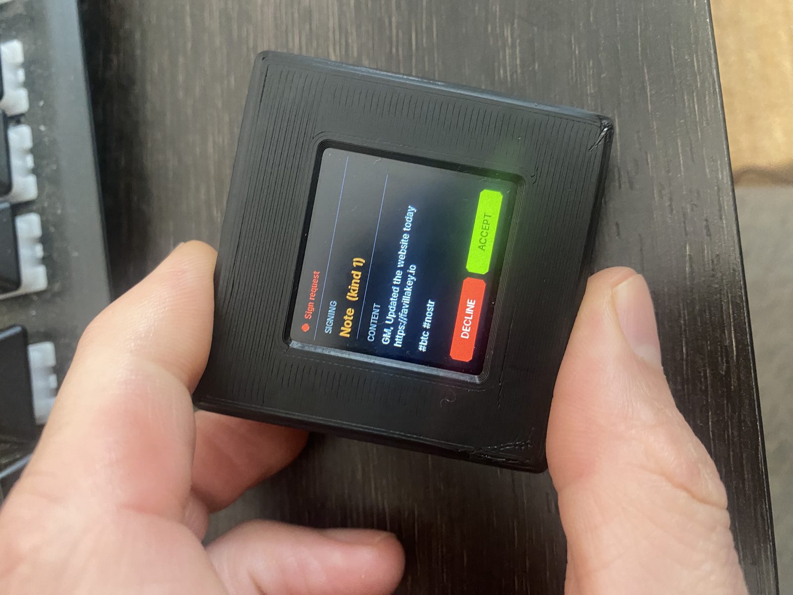 Favilla KEY prototype held in hand showing a Sign request screen with a real Nostr note (kind 1) ready to accept or decline